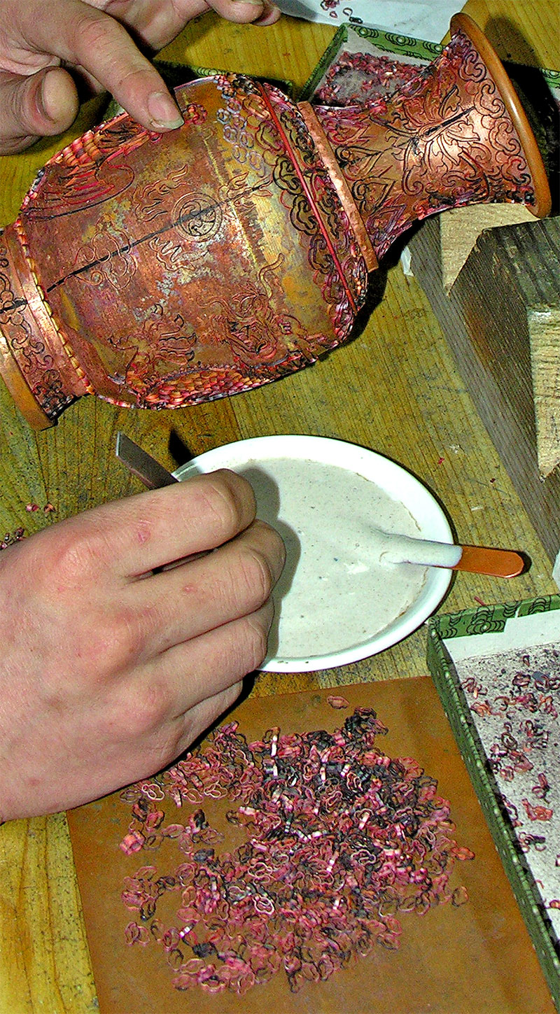 Un artesano moldea la filigrana de cobre y la adhiere a la plantilla del dibujo sobre la vasija, para hacer los moldes que contendrán el esmalte de colores. Foto: Dennis G. Jarvis - China-6474. CC BY-SA 2.0. Wikimedia para «Cloisonné_of_China».