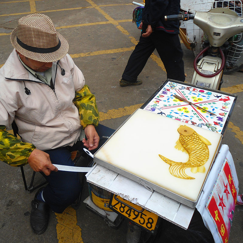 Mesa de trabajo callejera del pintor de azúcar en el parque de la Montaña del Toro Dorado, en Haiku, provincia de Hanan. La ruleta marca el dibujo al azar y el pintor lo ejecuta con el azúcar caliente.Foto de Anna Frodesiak, Wikimedia commons, dominio público